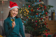 Happy Asian woman wears santa hat celebrating and toasting a glass of champagne with christmas tree in background