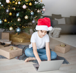 A boy sits near a Christmas tree with a laptop.