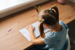 © dikushin - High-angle view of unrecognizable little child girl learning writing doing homework sitting at home table by window. Top view of primary schoolgirl child studying alone making note with pen at bedroom