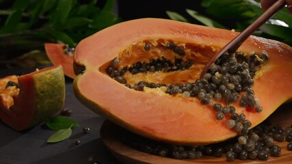 Wall Mural - Half papaya fruit with seeds on a vintage table with leaves of tropical plants. A hand with a wooden spoon cleans the papaya fruit from the seeds. Horizontal slow movement of the camera from left to r