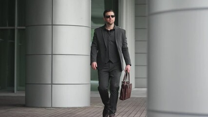 Wall Mural - Slow motion of cool businessman in suit with sunglasses holding briefcase walking in the office building at urban city confidence man 