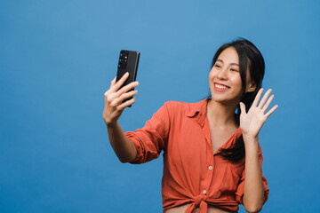 Wall Mural - Smiling adorable Asian female making selfie photo on smart phone with positive expression in casual clothing and stand isolated on blue background. Happy adorable glad woman rejoices success.