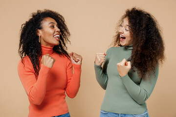 Canvas Print - Two fun young curly black women girlfriends 20s wearing casual shirts clothes look at each other do winner gesture celebrate clenching fists isolated on plain pastel beige background studio portrait.