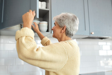 Canvas Print - leisure, household and people concept - happy smiling senior woman opening kitchen locker with crockery at home