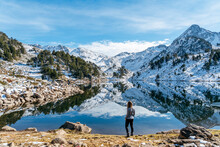 Woman And Reflection Of The Snowy Mountains In The Beautiful Baciver Lake In The Pyrenees Mountains Of Val D'Aran (Aran Valley), Lleida, Catalonia, Spain