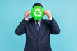© khosrork - Unknown male person wearing official style suit hiding face behind green recycling sign, garbage sorting and environment protection, thinking green. Indoor studio shot isolated on blue background.