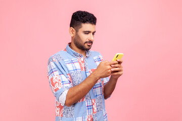 Side view of serious concentrated brunette man with beard holding cell phone in hands, chatting or checking e-mail, gadget addiction. Indoor studio shot isolated on pink background.