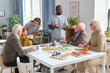 © pressmaster - Group of senior people playing board game at the table and talking to medical staff in the room of nursing home