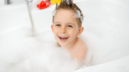 Wall Mural - Portrait of cheerful laughing boy washing in bath with foam suds and foam