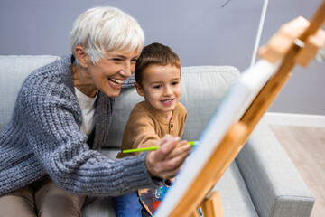 Grandson and grandma painting on canvas. Artist teaching young boy how to paint at home. Happy grandmother and a little boy painting. Senior woman with child painting on canvas.