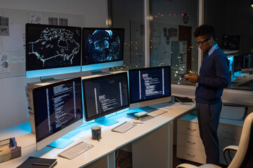 young african programmer texting in smartphone while standing by workplace against large office wind