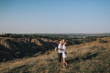 A loving groom and a beautiful blonde bride in a white dress tenderly hug in nature on the background of hills and cliffs in autumn in sunny weather. Wedding photo of smiling newlyweds.