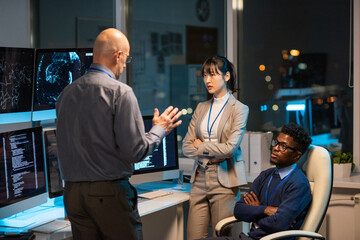 Poster - Mature manager giving instructions to young intercultural subordinates at working meeting late at night