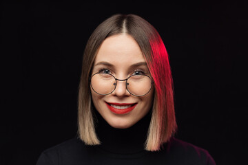 Close up woman portrait wearing glasses with black frames, with red lips, looking to camera on black background and pink light on hair with focused face expression