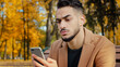 © Yuliia - Portrait young serious guy use app hispanic man looking at phone screen browses news on telephone concentrated male sits on bench in autumn park read using device businessman holds smartphone outdoors
