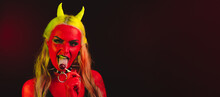Red Demon Girl Whit Yellow Eyes And Yellow Horns And Hair Wearing Necklaces With Spikes On Dark Background Looking At The Camera Holding A Red Lollipop On Her Tongue