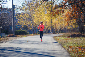 Wall Mural - Young fitness woman running on the road in the cold morning in winter. Sporty girl in orange long-sleeve jogging in the sunny morning