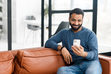Attractive Indian bearded man with a pleasant smile chilling and sitting on the couch, chatting with friend in social media on mobile phone, dating, flirting, spending leisure time online