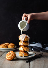 Hand Pouring Icing On Fresh Cinnamon Buns Stacked High On A Plate.