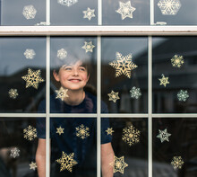 Happy Boy Looking Out Through A Window Covered In Snowflake Decals.