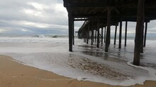 Walking Backing Up Under A Wooden Pier Along The Coast As Large Sea Ocean Salt Water Waves From Tropical Storm Surge Hurricane Season Crash Onto The Beach Sandy Shore Ion A Cloudy And Windy