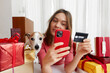 Woman buys gifts online through a smartphone with her pet dog sitting at a table among gifts packed boxes in colored paper with bows and ribbons at home