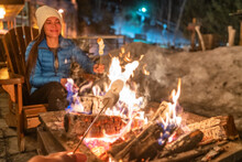 Winter Campfire Couple Roasting Marshmallows In Firepit For Smores At Ski Holiday Resort. Asian Happy Woman Relaxing After Ski.