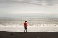 Boy In Red Sweater Looking At Ocean On Black Lava Beach Of Vik, Iceland At Sun Set