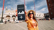 Audio tour online app on digital mobile smartphone. Happy young student woman holding phone listening audioguide at San Marco square in Venice, Italy. Simultaneous translation devices.