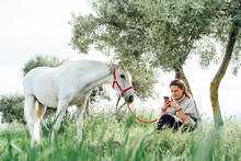 Young Woman Using Smart Phone While Sitting Under Tree By Horse At Field