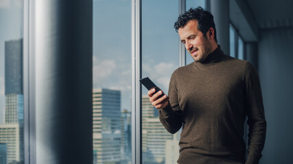 Poster - Thoughtful Arab Businessman Using Smartphone Standing in Office Looking out of the Window on Big City. Charismatic Digital Entrepreneur Plan Investment Strategy for e-Commerce Startup.