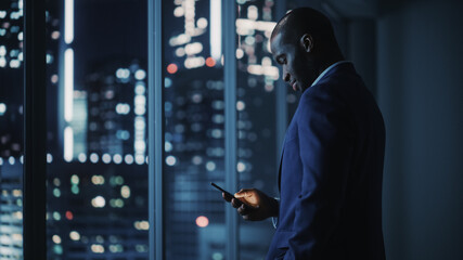 Poster - Night Office: Successful Black Businessman Wearing Suit Standing, Using Smartphone Looking out of the Window on a Big City. African-American CEO Thinking of Investment Strategy in e-Commerce Software
