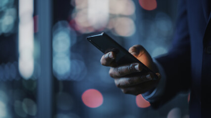 Poster - Night Office: Successful Black Businessman Wearing Suit Standing, Using Smartphone. CEO Browsing Internet, Managing Social Media Strategy in e-Commerce Software. Focus on Hand with Mobile Phone