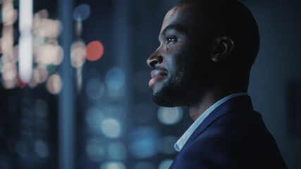 Wall Mural - Night Office: Stylish Close-up Portrait of Powerful Black Businessman Wearing Suit Standing, Looking out of the Window on a Big City. Ambitious African CEO Thinking of e-Commerce Investment