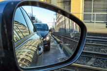 Car In The Rear View Mirror Next To Tram Rails . Reflections From Structured Buildings And Straight Lines Crossing . Tracking View . Car Followed By Stranger Or Detective .