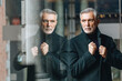 © Westend61 - Mature man adjusting jacket while standing by transparent glass wall
