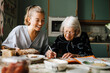 © Maskot - Senior woman solving crossword puzzle in book sitting by smiling nurse in kitchen at home
