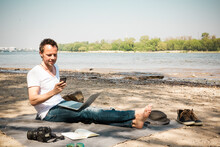Man Sitting On Blanket At A River Using Laptop And Cell Phone