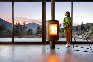 Woman enjoy great view on mountains while sitting near fireplace at modern living room at sunset . Concept of rest in houses or cabins on nature. Solitude in nature and escape from everyday life