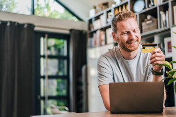 Wall Mural - Happy cheerful smiling young adult man doing online shopping or e-shopping satisfied entrepreneur making online payment paying for service or goods happy spending free time