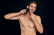 Handsome young bearded man isolated. Portrait of topless muscular man is standing on black background with electric Shaver in hand while shaving. Men care concept