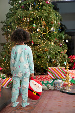 Little Girl In Onesie Pajamas Standing And Looking At Christmas Tree In Wonderment. 