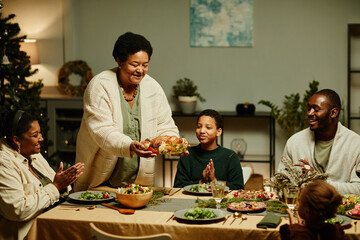 Wall Mural - Portrait of caring African-American grandmother bringing food to table while celebrating Thanksgiving with big happy family