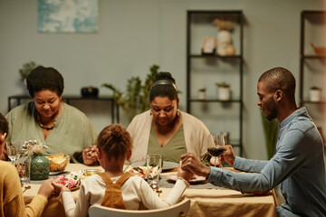 Canvas Print - Portrait of modern African-American family holding hands while saying grace during dinner party at home