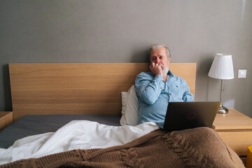 Wall Mural - Thoughtful gray-haired senior male with beard using laptop computer lying sitting on comfortable bed at home. Handsome bearded mature older man in pajamas having online video call via notebook.