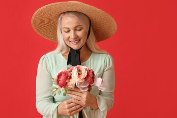 Wall Mural - Beautiful mature woman in straw hat holding bouquet of flowers on red background