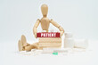 © Dzmitry - A figurine of a man sitting among pills lifts a red wooden block with an inscription PATIENT from a wall of blocks.
