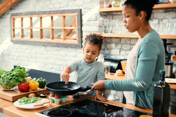 Wall Mural - Black little girl and her mother prepare food together in kitchen.