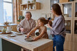 © Tatiana Timofeeva/Stocksy - mom braids her daughter's braid while grandma cooks lunch