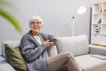 Beautiful 50 Years Old Caucasian Lady Sit Down On The Sofa Drinking Tea And Reading A Book For Afternoon Indoor Leisure Activity At Home - After Work Lifestyle For People Concept